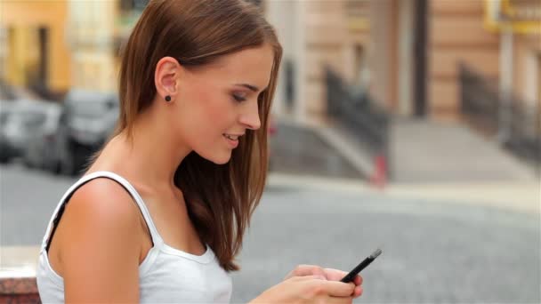 Vista lateral de una mujer de moda usando un teléfono inteligente sentado en un banco en la calle
 - Metraje, vídeo
