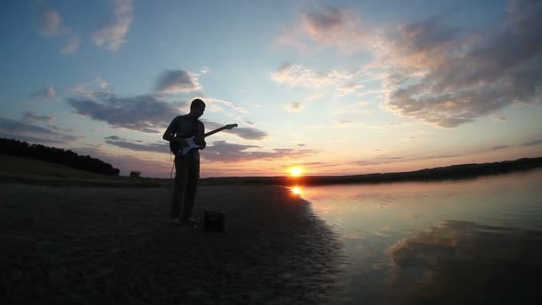 ο άνθρωπος σε εξωτερικούς χώρους κατά το ηλιοβασίλεμα, κοντά στη λίμνη παίζει ηλεκτρική κιθάρα βράδυ ήλιος δύει βίντεο - Πλάνα, βίντεο