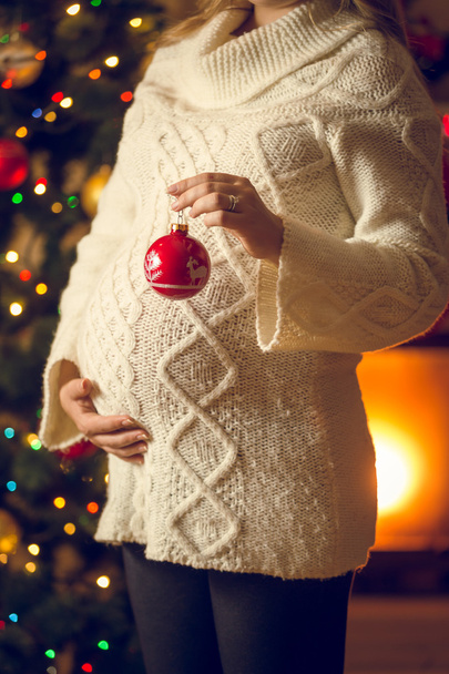 έγκυος γυναίκα στο μαλλί πουλόβερ κρατώντας κόκκινη Χριστουγεννιάτικη μπάλα - Φωτογραφία, εικόνα