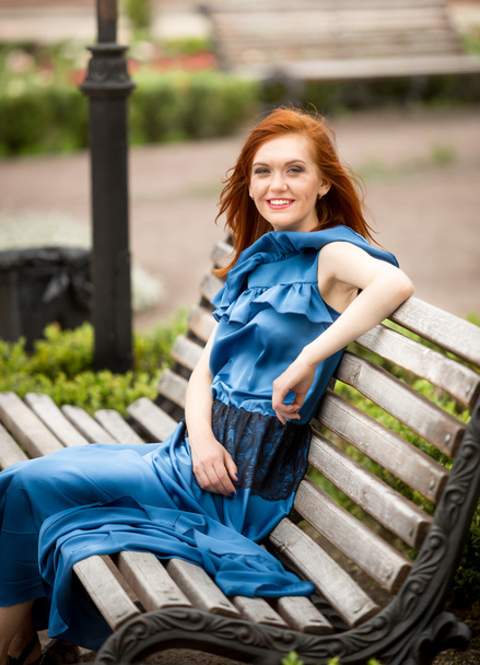 redhead woman in blue dress sitting on wooden bench at park - Valokuva, kuva