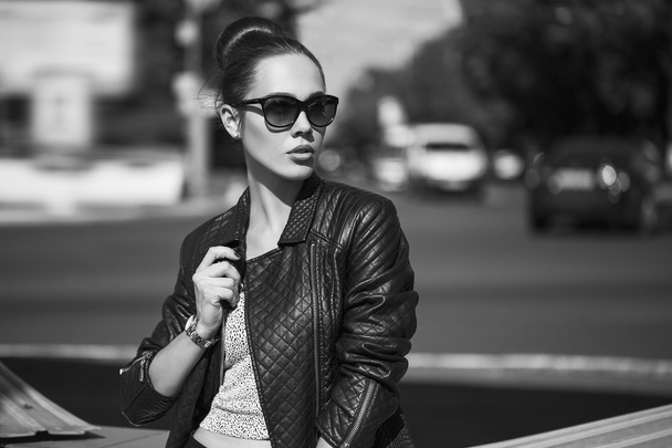 Female beauty concept. Portrait of fashionable young girl in casual black jeans, black jacket, white crop-top, sunglasses and small  bag posing on the street. Perfect hair & skin. Vogue style. outdoor shot - 写真・画像