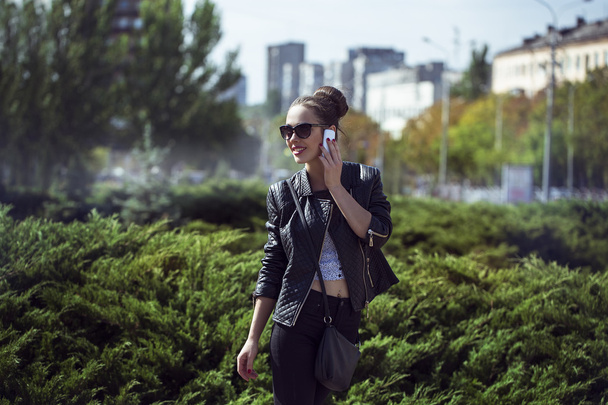 Concepto de belleza femenina. Retrato de una joven de moda en jeans negros casuales, chaqueta negra, top blanco, gafas de sol y bolsita posando en la calle. Cabello y piel perfectos. Estilo Vogue. tiro al aire libre
 - Foto, Imagen