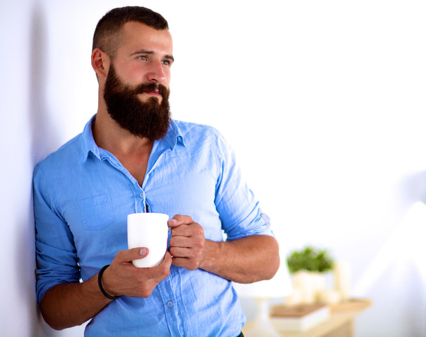 Young man  standing near wall and holding cup of coffee in office - Foto, Bild