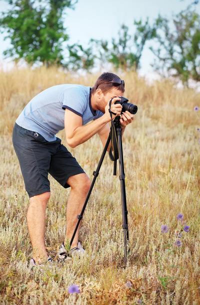Mies kolmijalka ja kamera seisoo niityllä ja tekee na
 - Valokuva, kuva