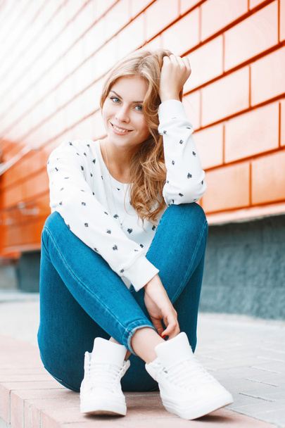 Portrait of a young beautiful girl in a white sweater, jeans and white sneakers sitting on a tile near the orange wall - Photo, Image
