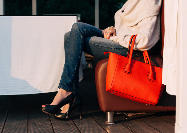 Girl sitting on the sofa of summer cafe with a big red super fashionable handbags in a sweater jeans and sneakers on a warm summer evening. warm colors - Foto, Imagem