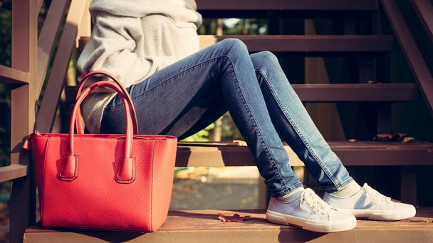 Girl sitting on the stairs with a big red super fashionable handbags in a sweater jeans and sneakers on a warm summer evening. Warm colors - Фото, изображение