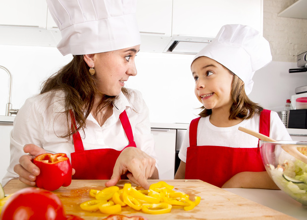 happy mother and little daughter at home kitchen preparing paprika salad in apron and cook hat - Fotó, kép