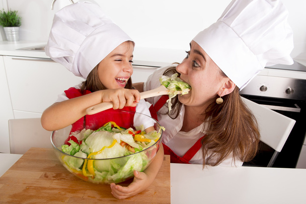 glückliche Mutter und kleine Tochter zu Hause Küche bereitet Salat in Schürze und Kochmütze - Foto, Bild