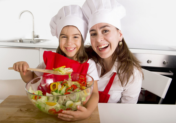 feliz madre e hija pequeña en la cocina casera preparando ensalada en delantal y sombrero de cocinero
 - Foto, Imagen