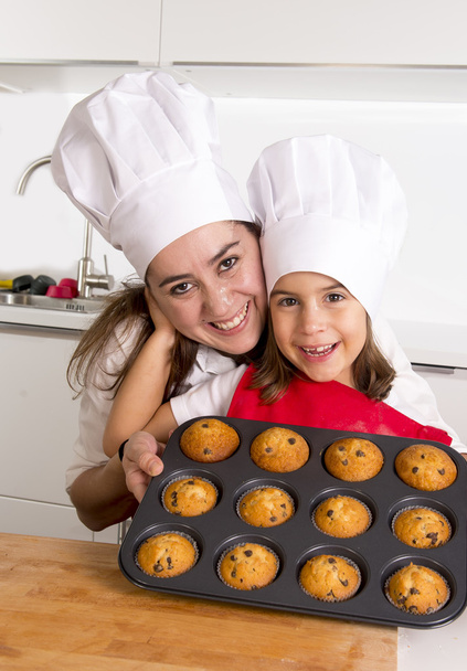 happy mother with daughter wearing apron and cook hat presenting muffin set baking together at home kitchen - Photo, Image