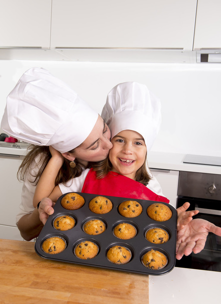 glückliche Mutter mit Tochter in Schürze und Kochmütze präsentiert Muffin-Set beim gemeinsamen Backen in der heimischen Küche - Foto, Bild