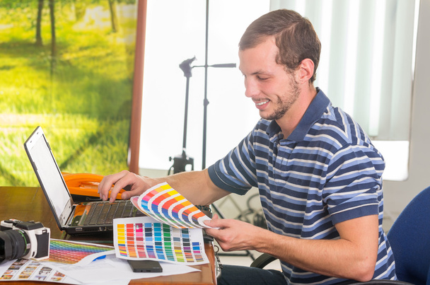 Man wearing blue white striped t-shirt sitting by work desk looking at pantone palette, colormap - 写真・画像