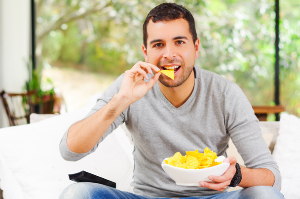 Hispanic male wearing light blue sweater plus denim jeans sitting in white sofa holding bowl of potato chips and remote control watching tv enthusiastically - Φωτογραφία, εικόνα