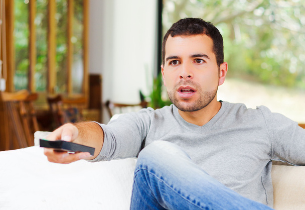 Hispanic male wearing light blue sweater plus denim jeans sitting in white sofa holding potato chip and remote control watching tv enthusiastically - Photo, image