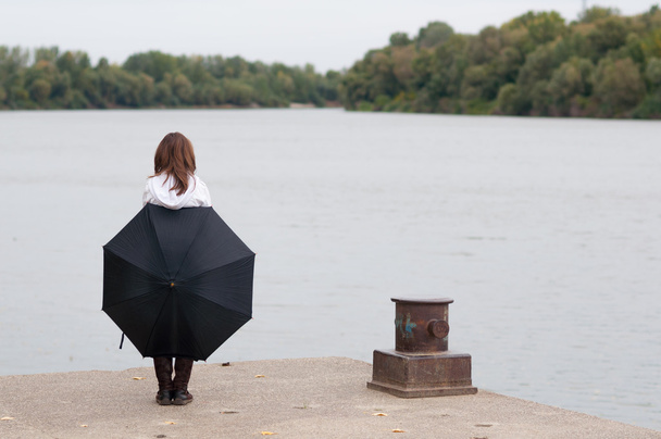 Lonely teenage girl with umbrella standing on river dock on gloomy autumn day - Photo, Image