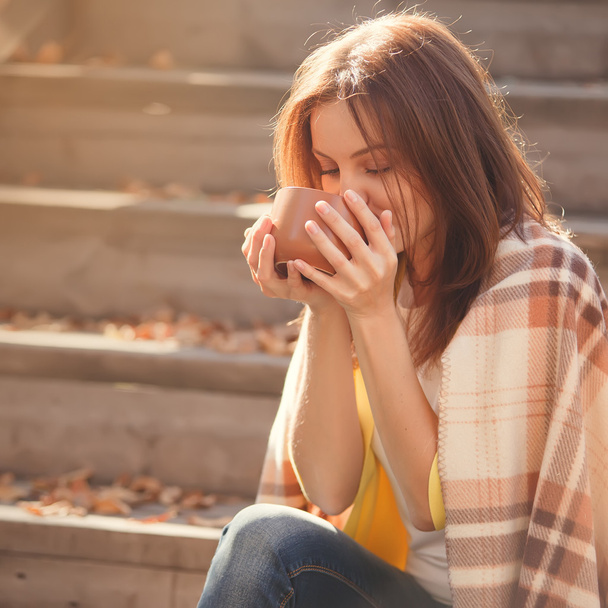 Jeune femme se reposant et buvant du thé assis dans le jardin d'automne sur les marches, enveloppé dans une couverture à carreaux de laine
. - Photo, image