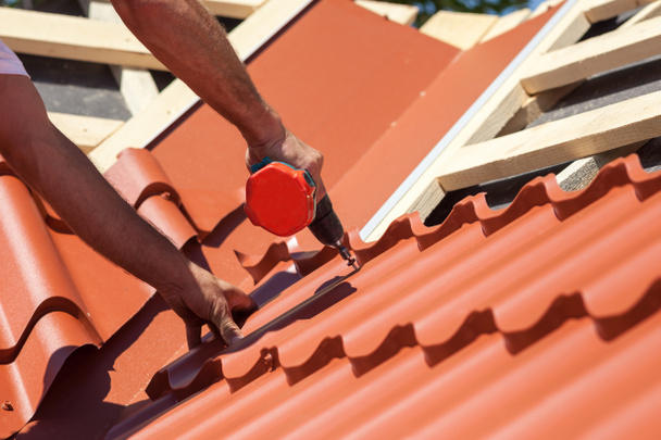 Trabajador en un techo con taladro eléctrico instalando azulejos de metal rojo en la casa de madera
 - Foto, Imagen