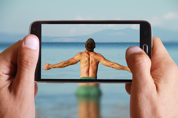 Male hand taking photo of Man Raising His Hands or Open arms standing back looking to sea blue sky horizon with cell, mobile phone. Strong muscular Men, perfect body, arms, back. Freedom Concept - Fotoğraf, Görsel