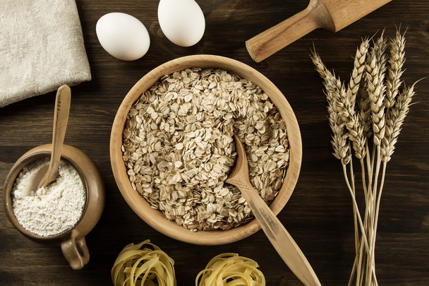 oatmeal flakes in a wooden bowl with a spoon, ears of wheat, pot of flour, kitchen utensils on the table. homemade, menu, recipe, mock up. - Foto, immagini