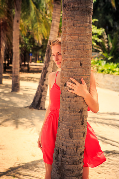 menina loira em vermelho olha para fora da palma olha para a frente contra as plantas
 - Foto, Imagem