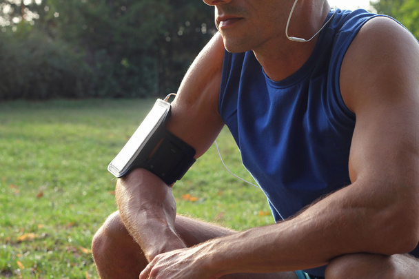 Male runner with mobile smart phone, listening to the music during workout. Athlete resting and relaxing after workout. Running, jogging, cardio, sport, active lifestyle concept - Photo, Image