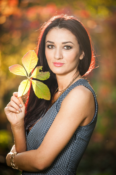 Beautiful woman in gray posing in autumnal park. Young brunette woman holding a leaf and spending time in autumn. Long dark hair woman smiling with a autumnal faded leaf in hand - Photo, Image