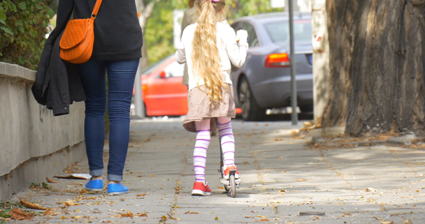 Äiti pieni blondi tyttö ratsastaa potkia skootteri katu nainen äiti kävelee pitkin tyttö tytär oranssi laukku ihmiset kävelevät pois pysäköity autot
 - Materiaali, video