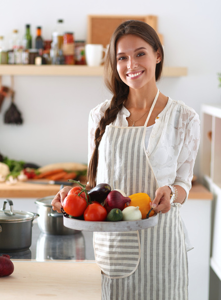 Smiling young woman holding vegetables standing in kitchen - Foto, afbeelding