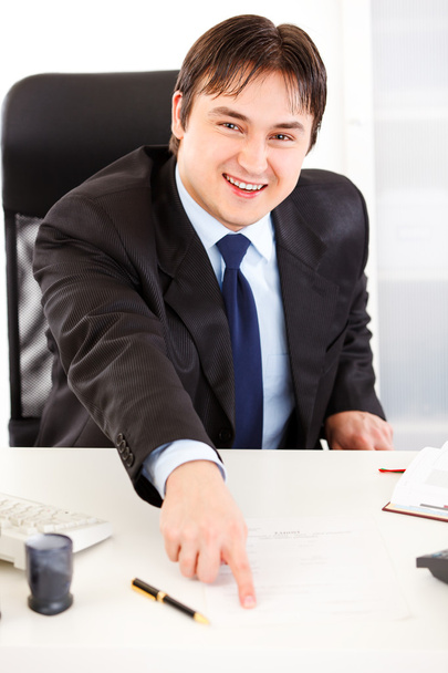 Smiling businessman sitting at office desk and pointing on document for si - Zdjęcie, obraz