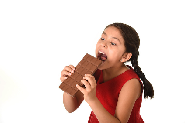Hispanic female girl wearing red dress holding with both hands big chocolate eating in happy excited face expression in sugary nutrition - Photo, image
