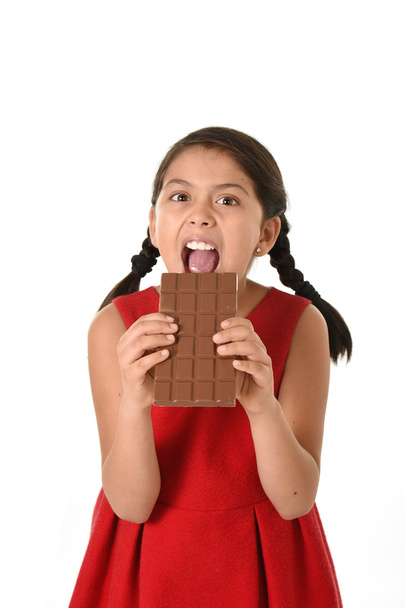 Hispanic female girl wearing red dress holding with both hands big chocolate eating in happy excited face expression in sugary nutrition - Фото, зображення