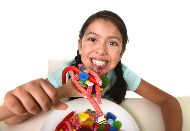 happy young girl holding spoon eating from dish full of candy lollipop and sugary things - Photo, Image
