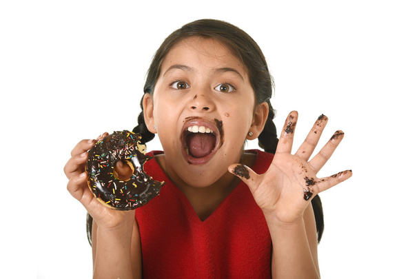 hispanic female child in red dress eating chocolate donut with hands and mouth stained and dirty smiling happy - Photo, Image