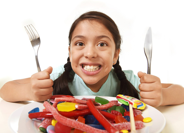 happy excited Latin female child holding fork and knife sitting at table ready for eat a dish full of candy - Photo, Image