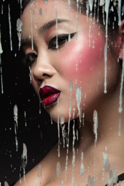 Beautiful Asian girl with bright make-up behind glass and drops of wax. Beauty face. - Photo, Image