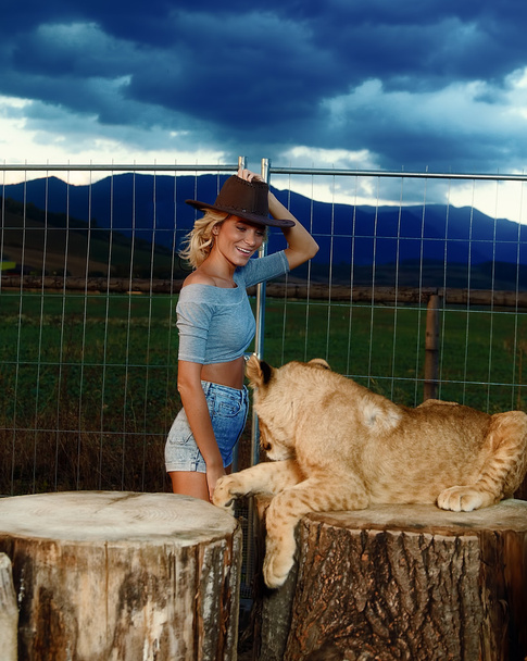 sexy woman playing with lion cub on background with beautiful sky and storm clouds. - Photo, Image