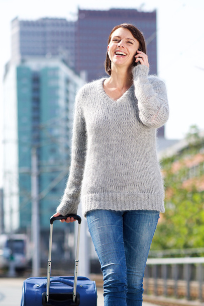 Mujer sonriente caminando y hablando por teléfono móvil con maleta
 - Foto, imagen
