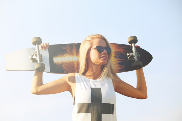 Young woman with skating board on blue sky background - Foto, Imagen