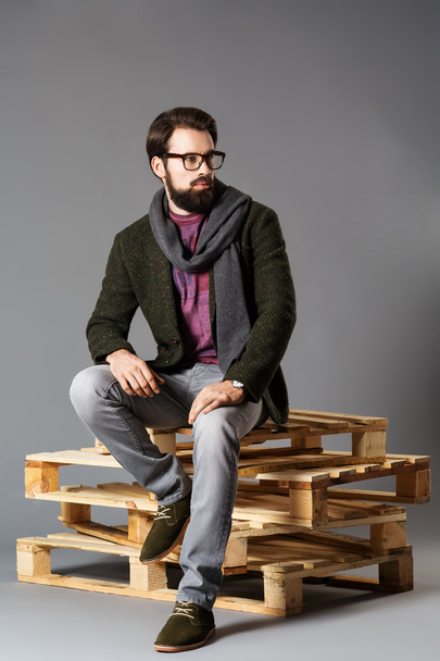 young stylish man with a beard, wearing a jacket sitting on pallets - Foto, Imagen