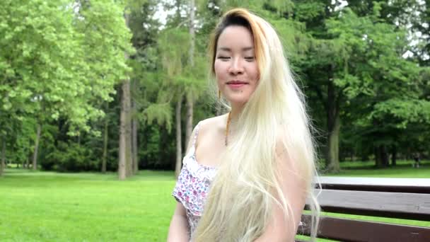 junge asiatische attraktive glückliche Frau sitzt auf der Bank im Park - lächelt in die Kamera - Nahaufnahme - Filmmaterial, Video