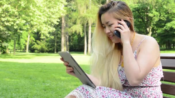 Genç Asyalı çekici gülümseyen kadın parkta bankta oturuyor - tabletle çalışıyor ve birileriyle konuşuyor - yakın plan - Video, Çekim