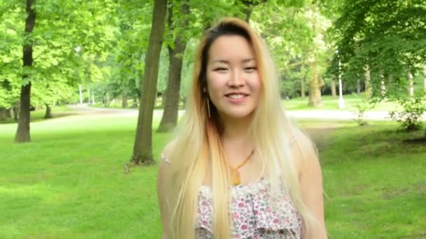 fiatal vonzó, boldog ázsiai nő fordul meg, és mosolyog a parkban - mosolyog a kamerába - Felvétel, videó