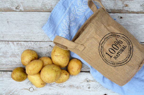 Organic fresh uncooked potatoes in a jute bag on a old weathered wooden white shelves background with vintage blue kitchen tea towel with empty copy space - Photo, Image