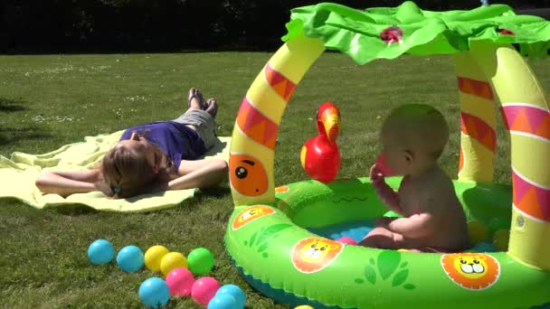 Careless mother lay on plaid and baby splash water in kiddie pool. 4K - Footage, Video