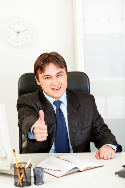 Pleased modern businessman sitting at office desk and showing thumbs up ges - Zdjęcie, obraz