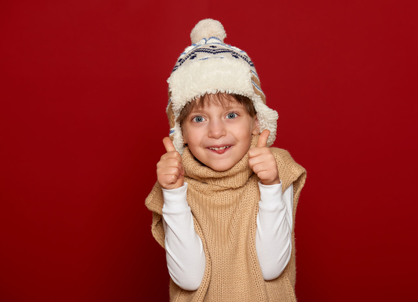 concepto de invierno - chica en sombrero y suéter muestran mejor gesto sobre fondo rojo
 - Foto, imagen