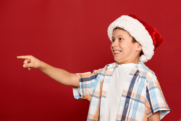 concepto de Navidad de vacaciones de invierno - niño en sombrero de santa señalar su dedo sobre el fondo rojo
 - Foto, imagen