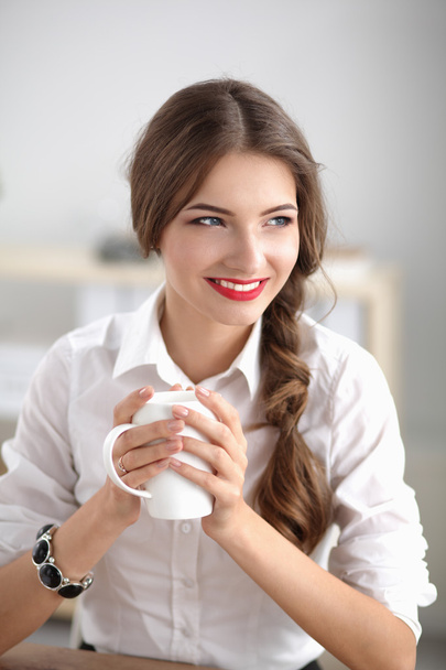 Young businesswoman sitting on the desk with cup in office - Foto, imagen