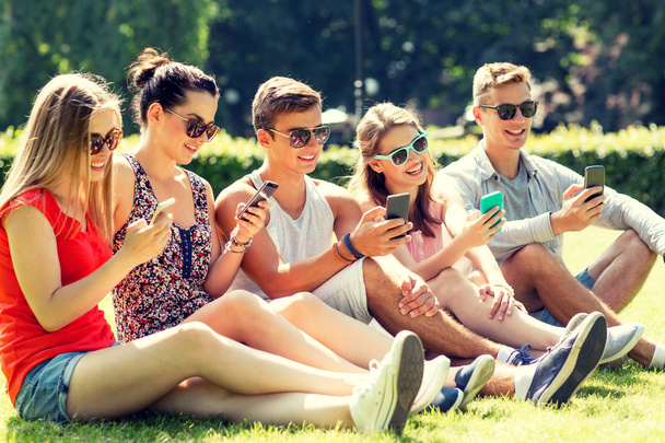 smiling friends with smartphones sitting on grass - Фото, изображение